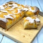 Slice of old fashioned Transylvanian peach cake with canned peaches served on a wooden cutting board.