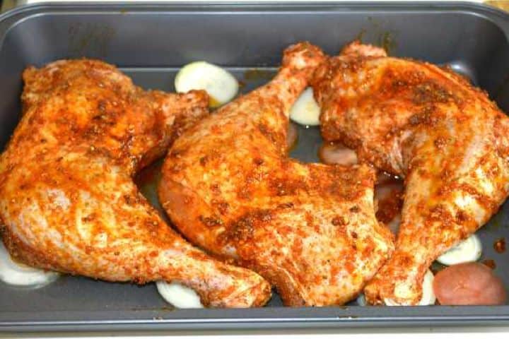 Seasoned turkey legs resting on a bed of coarsely sliced onions and tomatoes inside a baking tray with halved green pepper and garlic.