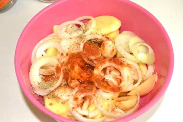 Tossing sliced potato rounds and white onion rings with sunflower oil, paprika, salt, and oregano in a bowl.