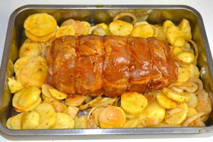 Tied stuffed pork loin arranged in a stainless steel baking tray surrounded by seasoned potatoes and onions.