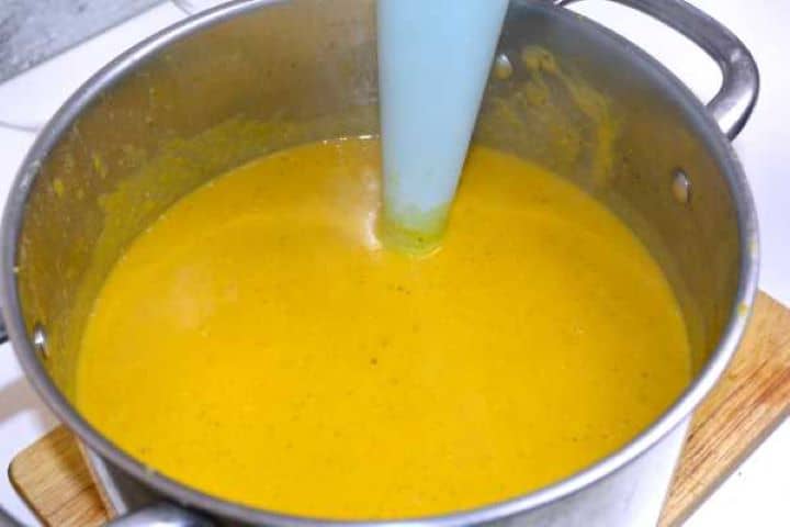 Pureeing cooked tender carrot and red lentil soup with a handheld immersion stick blender until smooth and creamy.