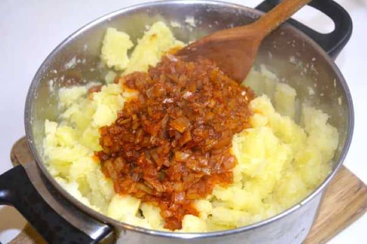 Adding warm sautéed paprika onions over rustic crushed potatoes in the cooking pot.