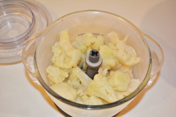 Boiled cauliflower florets and chopped leeks inside an electric food chopper.