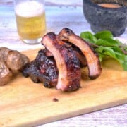 Sticky honey glazed pork ribs sliced on a rustic wooden cutting board with a cold beer and roasted potatoes.