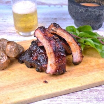 Sticky honey glazed pork ribs sliced on a rustic wooden cutting board with a cold beer and roasted potatoes.