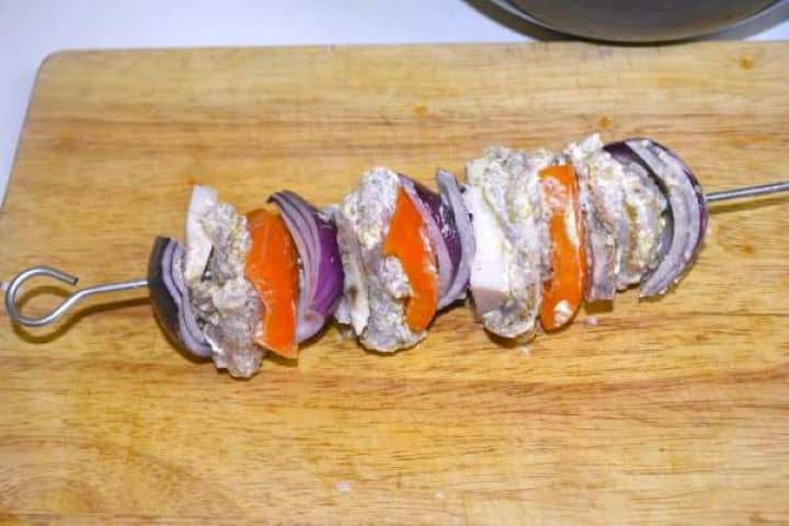 Raw chicken thigh kebabs layered with bell peppers, onions, and smoked fat on metal skewers.