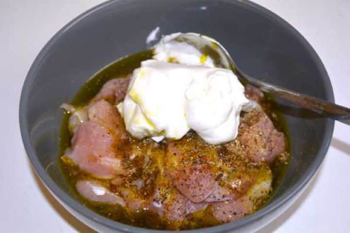 A large ceramic bowl containing bite-sized chicken thigh pieces well-coated in a thick, creamy Greek yogurt and spice marinade, ready for marinating.