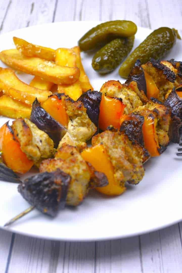 A detailed close-up of a serving of finished oven cooked chicken kebabs on wooden skewers, plated alongside a side of crispy french fries and mixed pickles, garnished with fresh parsley.