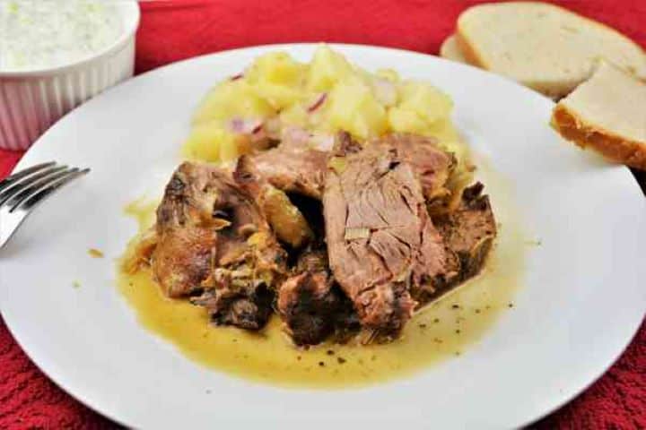Slow Roasted Leg of Lamb Recipe-Served on Plate With Potato Salad and Tzatziki Slow-roasted Greek leg of lamb served with potato salad, bread, and fresh tzatziki on a white plate.