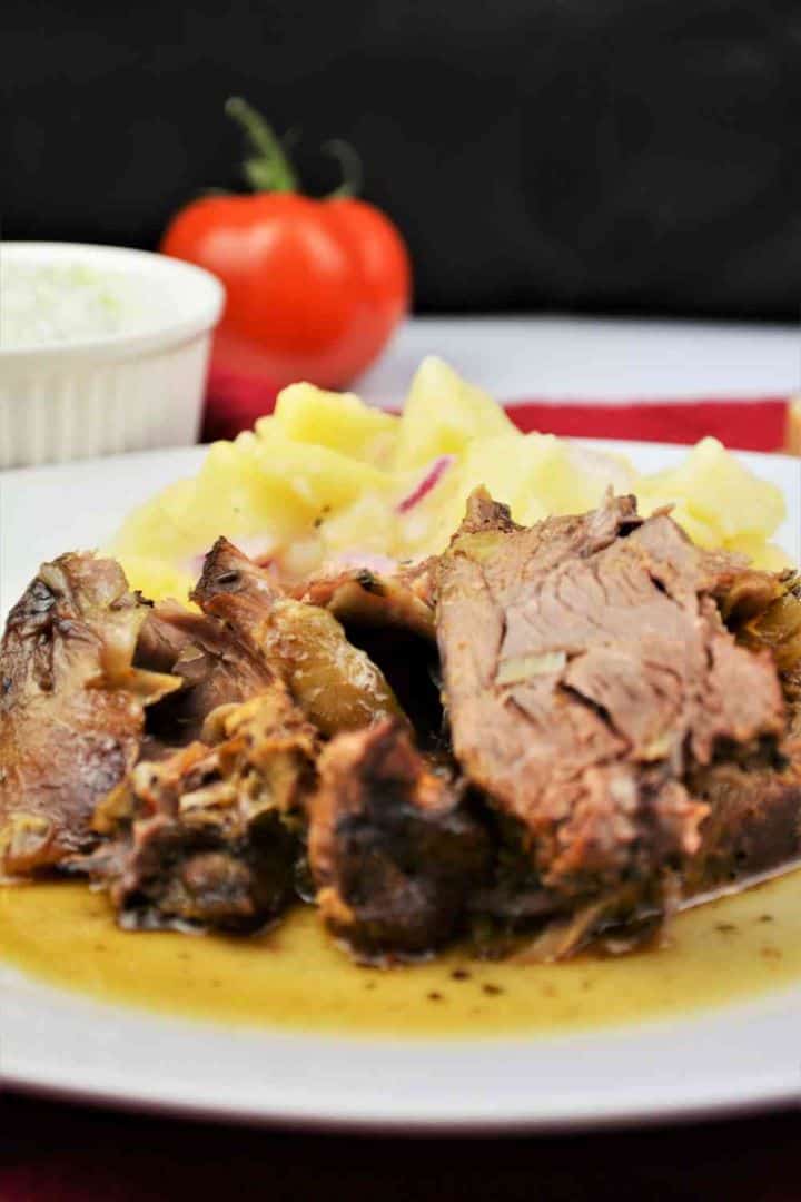 Slow-roasted Greek leg of lamb served with potato salad and fresh tzatziki on a white plate.
