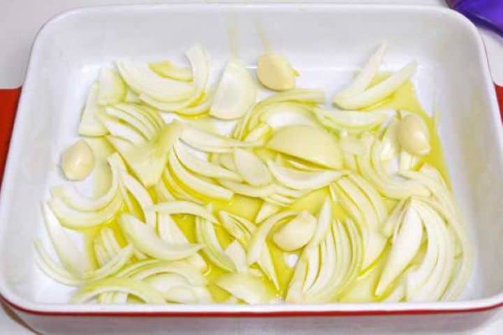 Sliced onions and smashed garlic arranged across the bottom of a ceramic baking dish.