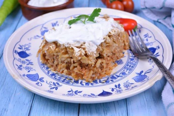 Layered Sauerkraut Casserole-Served on Plate With Sour Cream on the Top Close-up of a layered sauerkraut casserole slice showing distinct layers of seasoned pork, rice, and cabbage.