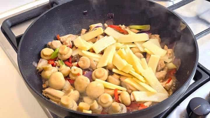 Combining chicken, red pepper, spring onions, button mushrooms, and bamboo shoots in a wok.