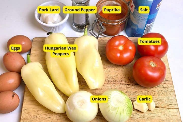 Fresh ingredients for Hungarian Lecsó including yellow wax peppers, ripe tomatoes, onions, garlic, eggs, and sweet paprika.