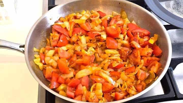 Adding thick tomato wedges to seasoned onions and peppers to form a rich vegetable stew base.