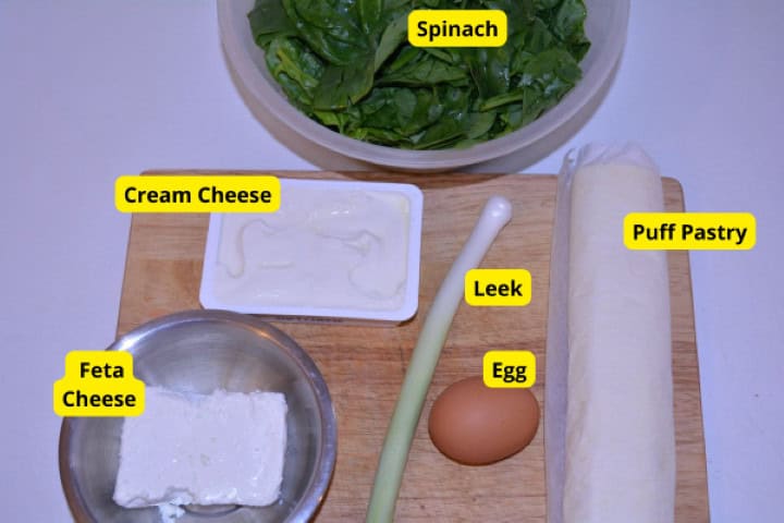 Ingredients for spinach and feta rolls including puff pastry, raw spinach, Greek feta, cream cheese, leek, and egg.