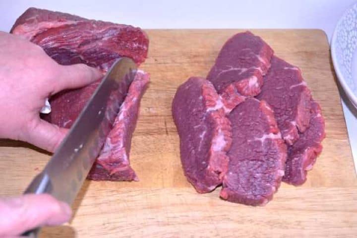 Meat Slicing raw beef roast into one-inch cutlets on a wooden cutting board.