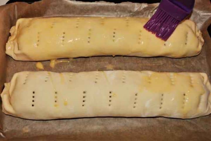 Apple Shortcrust Pastry Recipe-Unction the Rolled Shortcrust Pastry in the Baking Tray A pastry brush applying an egg wash glaze over a raw apple shortcrust pastry roll before baking.