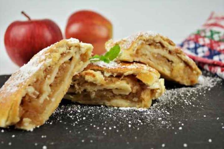 Apple Shortcrust Pastry Recipe-Served on the Platter With Icing Sugar Close-up of a warm slice of apple shortcrust pastry roll on a black platter with a vanilla icing sugar.