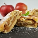 Golden baked apple shortcrust pastry roll sliced on a black platter dusted with icing sugar on top.