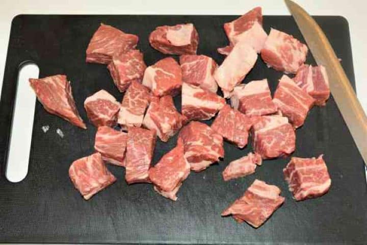 Dutch Oven Beef Stew-Beef Braising Steak Cut in Cubes A raw piece of marbled chuck roast cut into uniform 1-inch beef braising steak cubes on a kitchen cutting board.