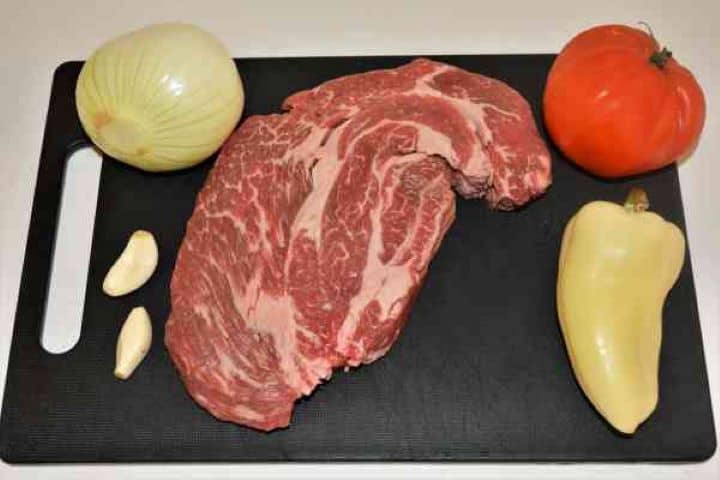 Dutch Oven Beef Stew-Beef, Onion, Yellow Pepper, Tomato and Garlic Cloves Raw beef braising steak, a whole white onion, yellow bell pepper, ripe tomato, and fresh garlic cloves arranged on a chopping board.