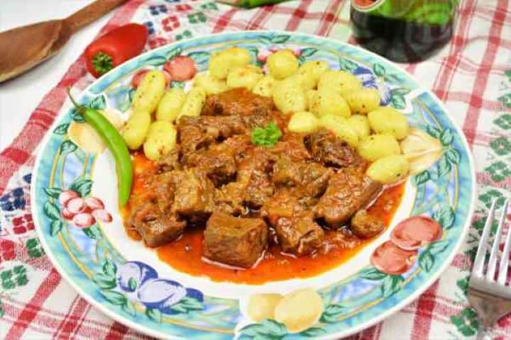 Dutch Oven Beef Stew-Served on Plate With Gnocchi and Chilli A colorful ceramic plate featuring authentic Hungarian style beef stew, potato gnocchi, and a green chili pepper side.