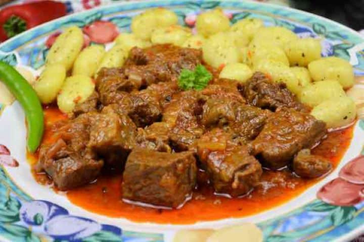 Dutch Oven Beef Stew-Served on Plate With Gnocchi and Chilli A close-up view of tender Dutch oven beef stew with paprika poured over boiled potato gnocchi on a colorful dinner plate.